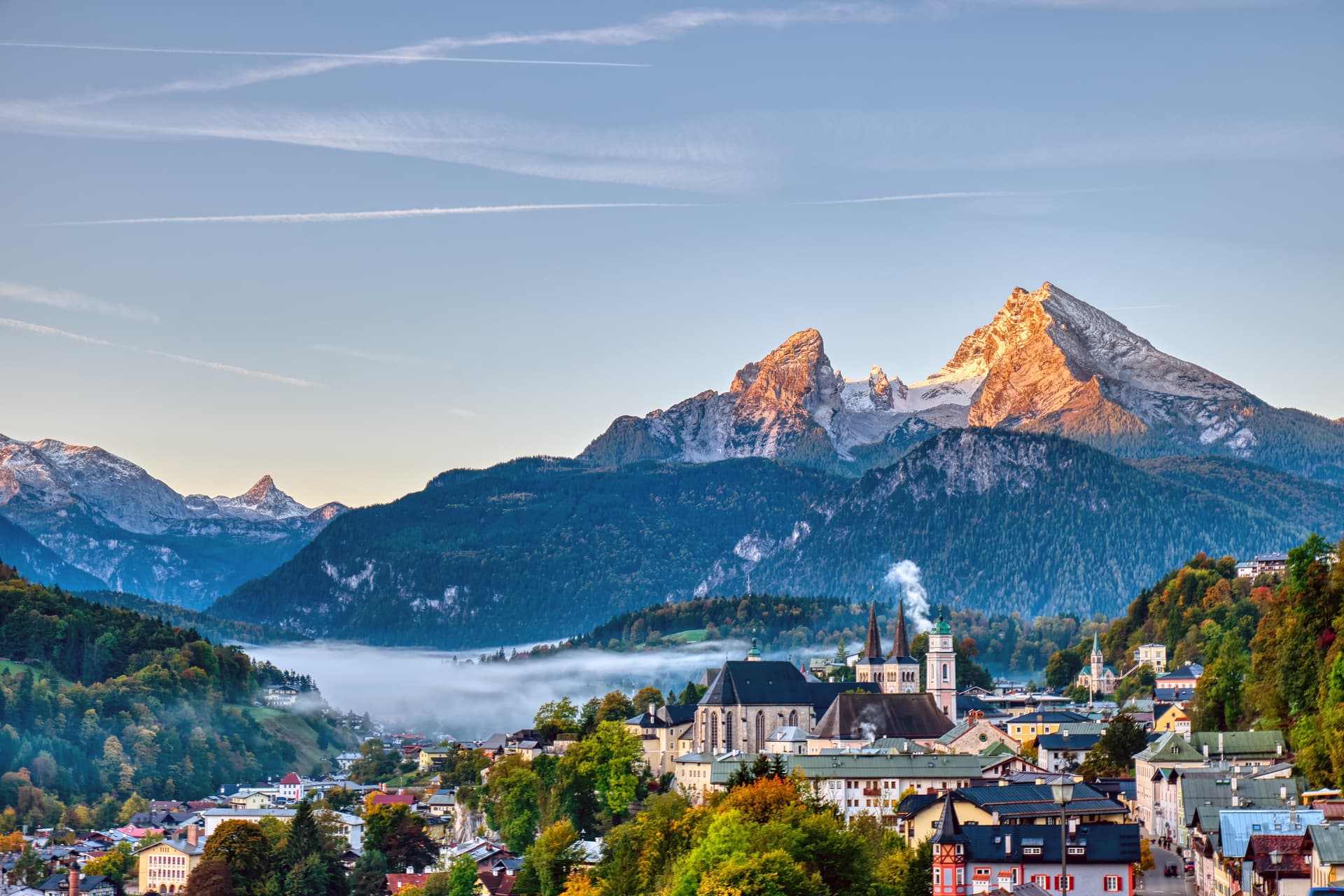Berchtesgaden town with Watzmann mountain peaks at sunrise, low fog, and autumn trees.