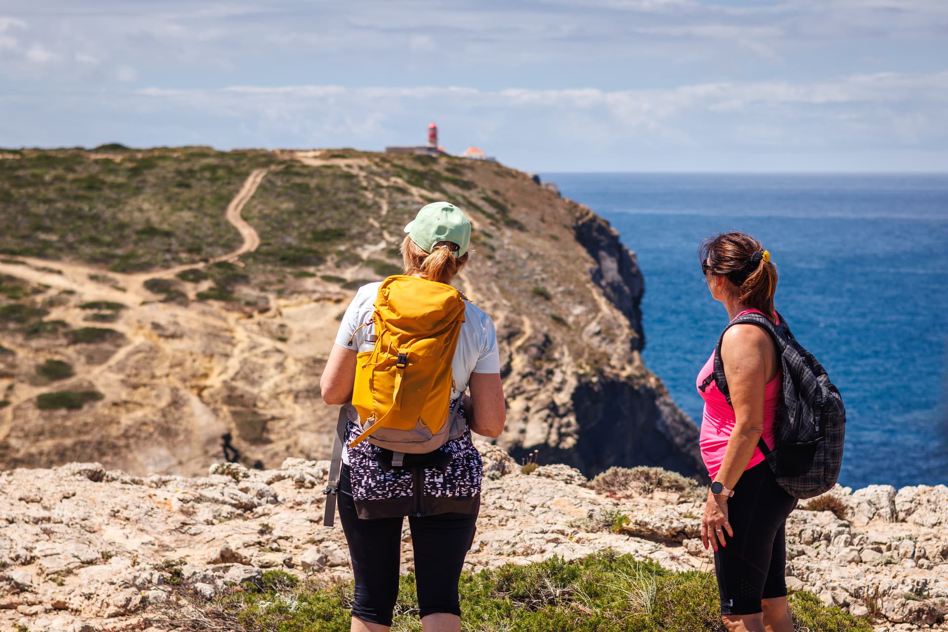 Hikers with backpacks on rocky trail overlooking the sea and a lighthouse in Algarve, Portugal.