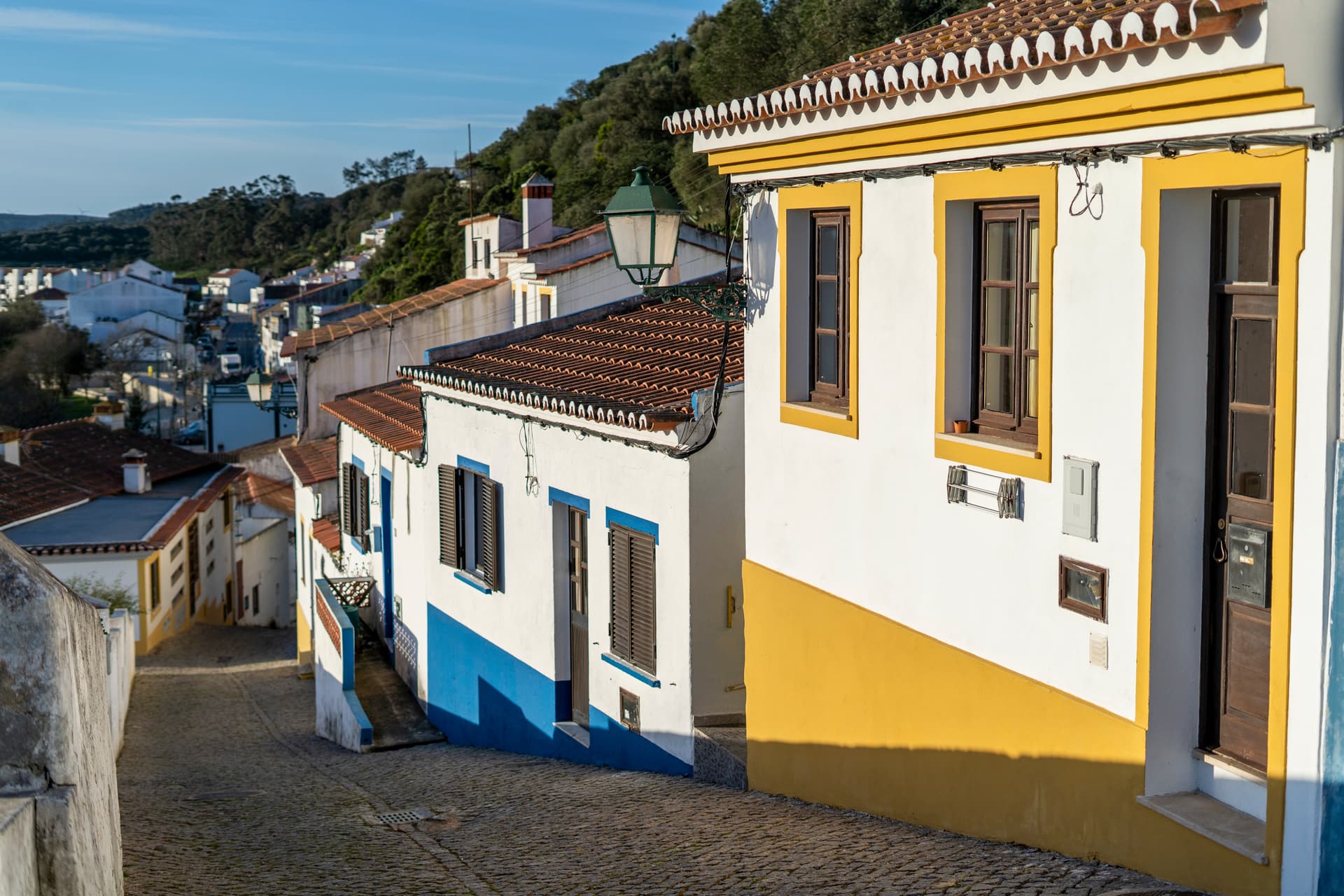 Cobblestone street sloping downhill past white houses with yellow and blue trim in Aljezur, Algarve, Portugal.