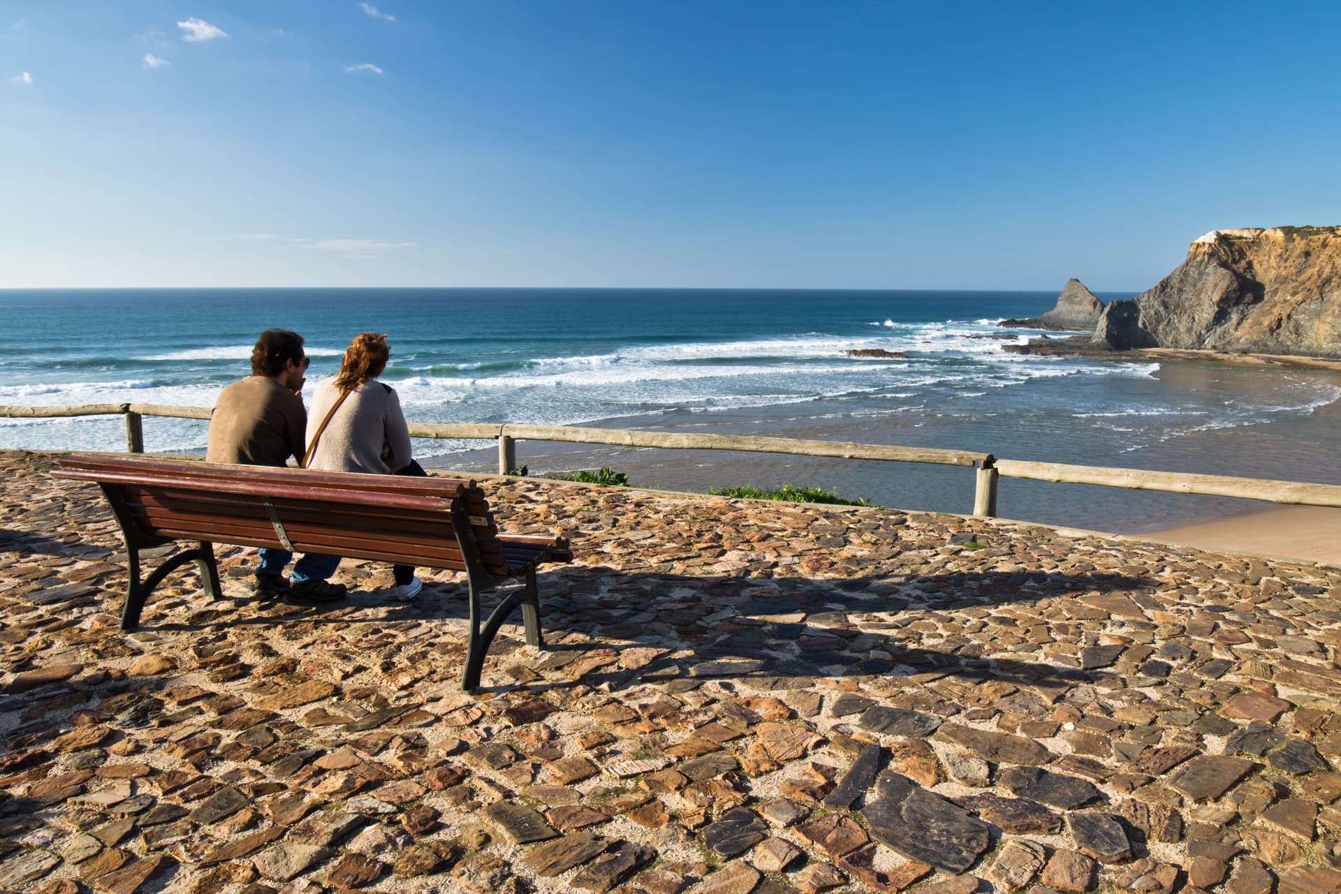 Couple sitting on bench overlooking waves crashing on Arrifana Beach with cliffs under blue sky.