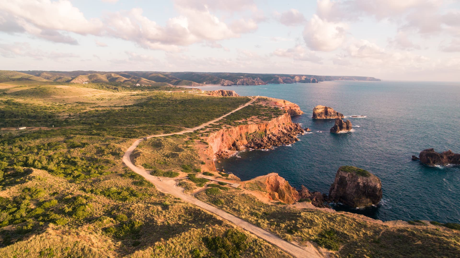 Coastal cliffs near Carrapateira, Portugal, with dirt road and sea stacks under cloudy sky.