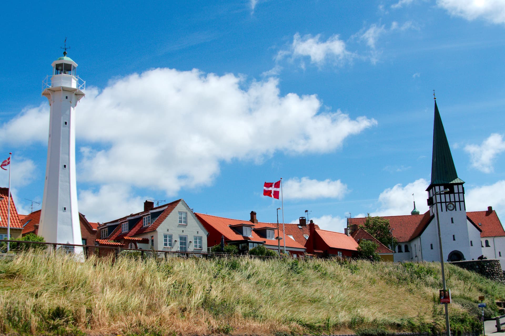 White lighthouse, church, and Danish flags above grassy dune in Rønne, Bornholm.