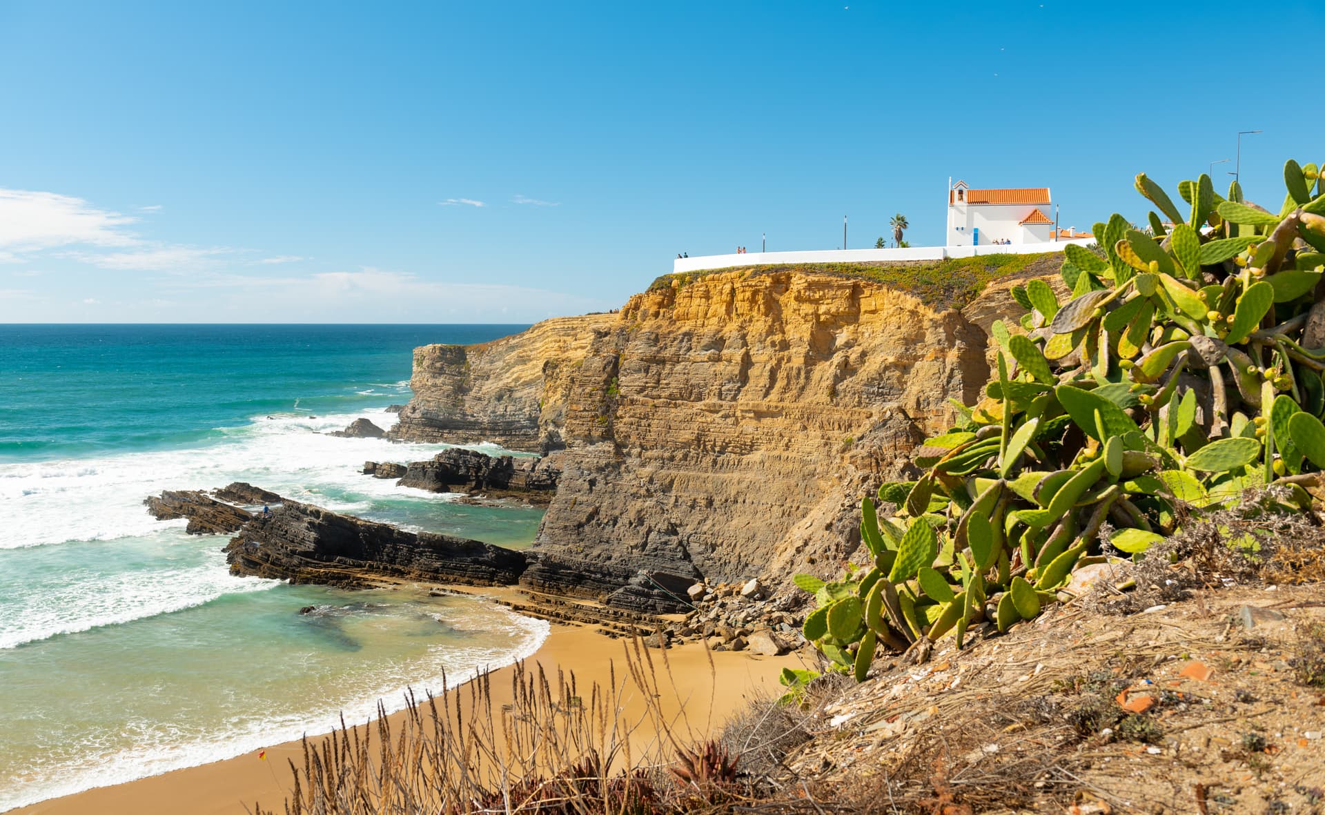 Zambujeira do Mar beach cliffs with white chapel, cactus, and turquoise autumn sea.