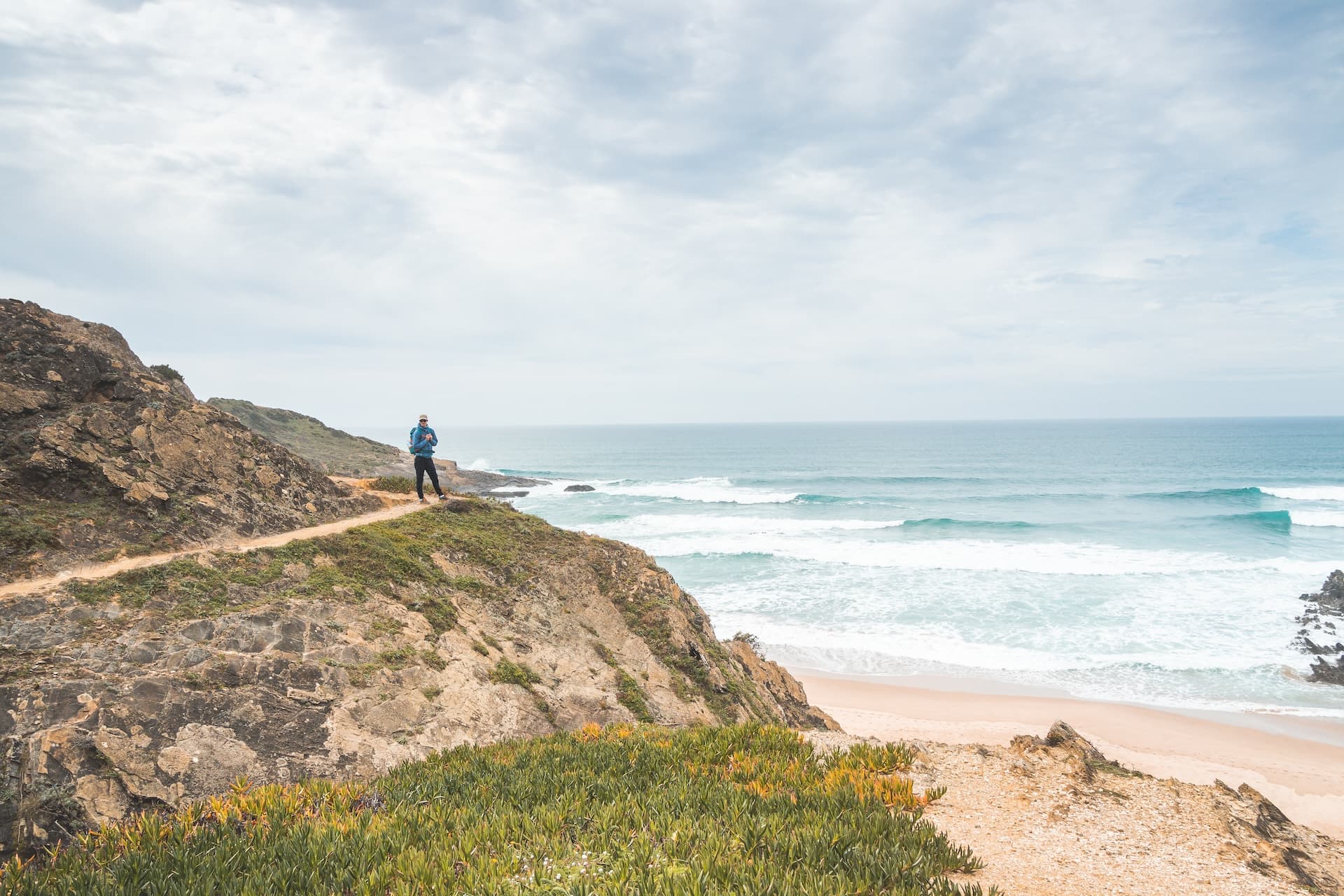 Hiker on cliff trail overlooking beach and Atlantic waves near Zambujeira do Mar.
