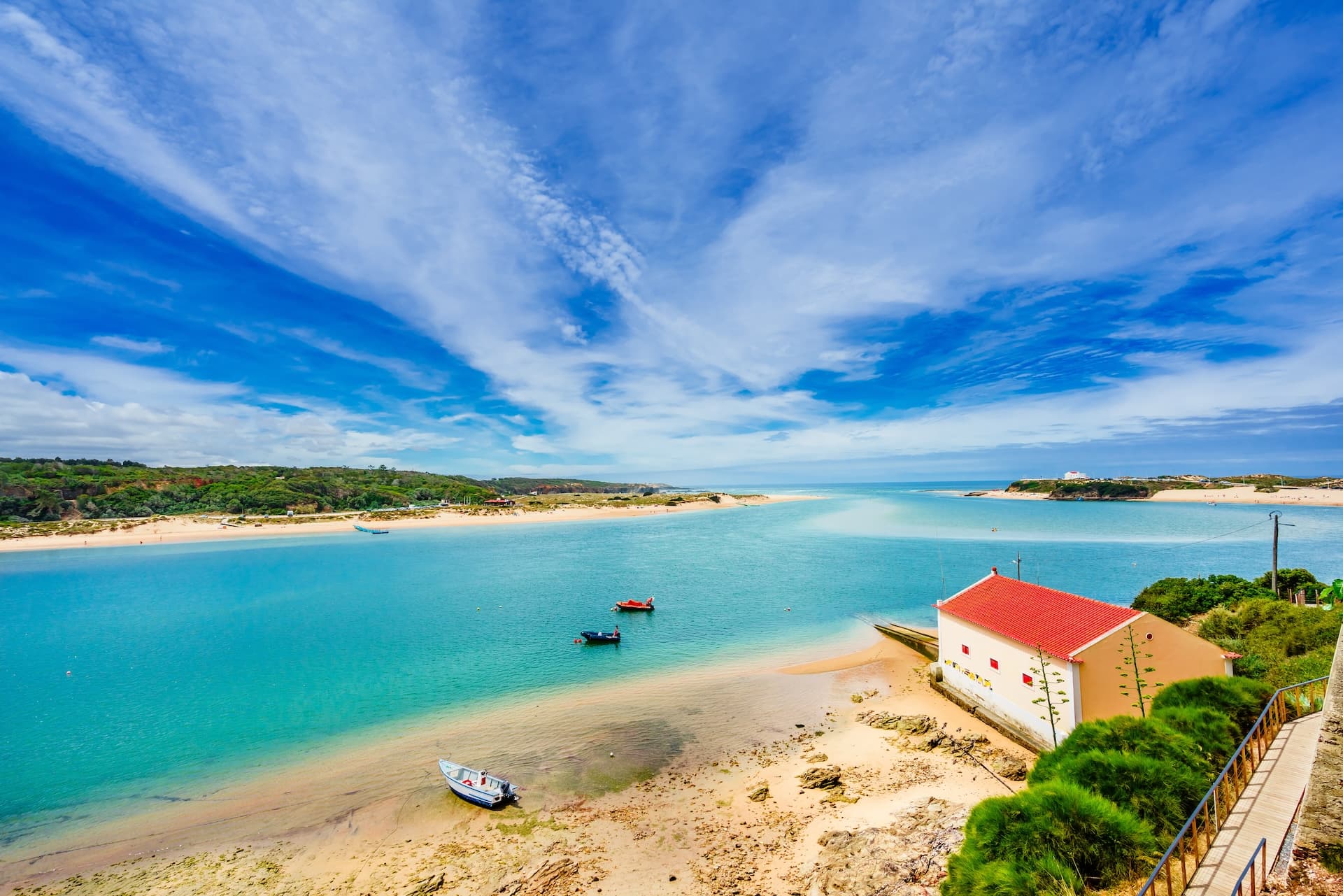 View on landscape with turquoise river, sandy shore, and white house with red roof in Vila Nova de Milfontes, Portugal.