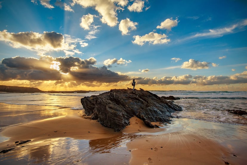 Person standing on large rock outcrop on sandy beach at sunset with sun rays through clouds.