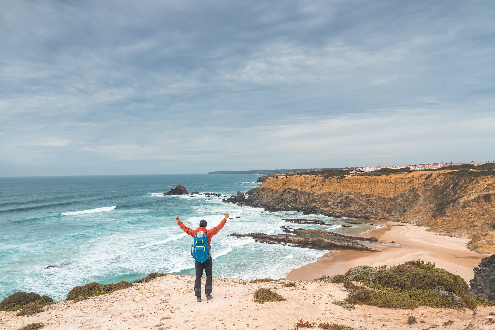 Hiker with arms raised overlooking beach cove and cliffs near Zambujeira do Mar, Odemira region.