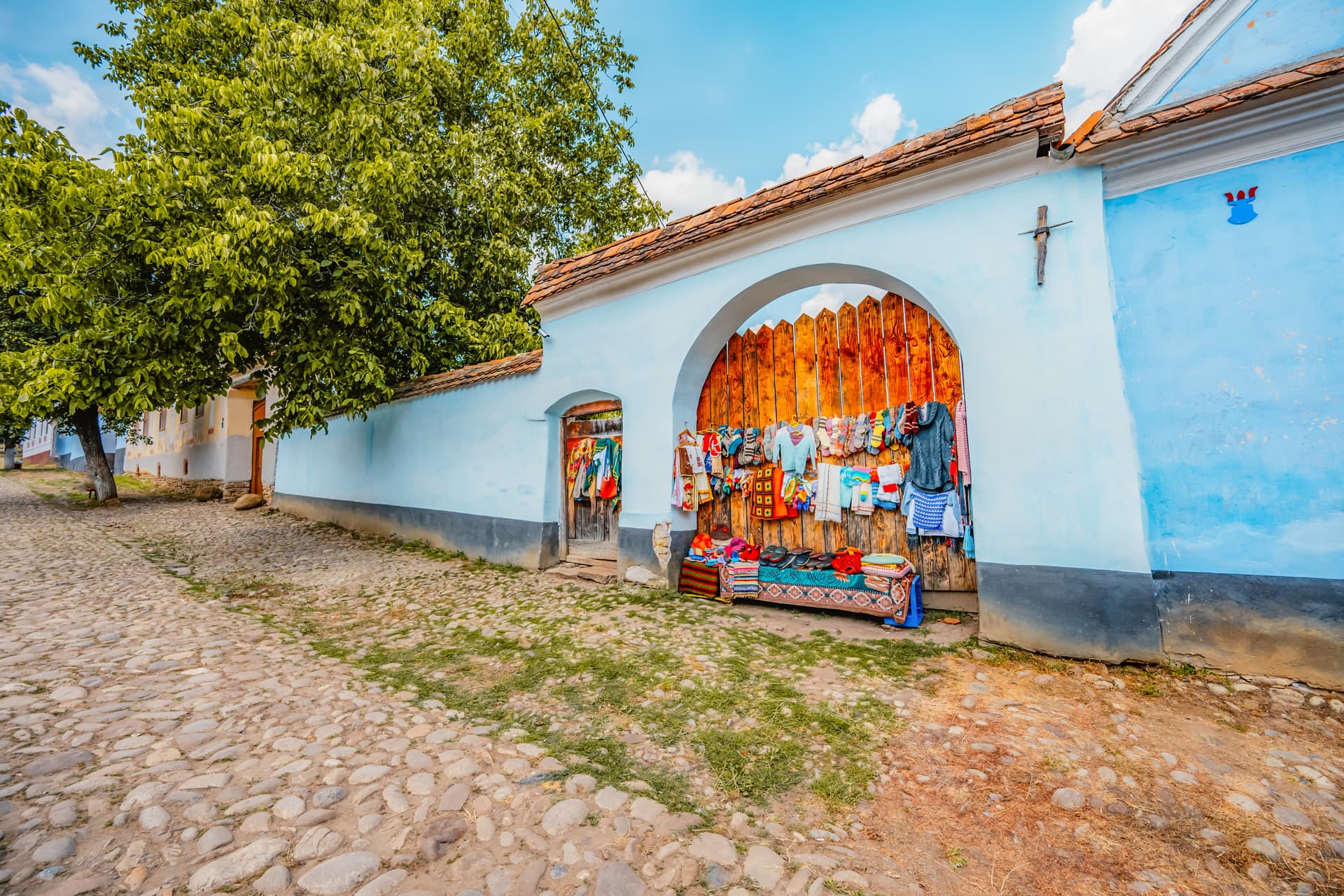 Street vendor selling colorful crafts and textiles outside a light blue wall with an arched wooden gate.