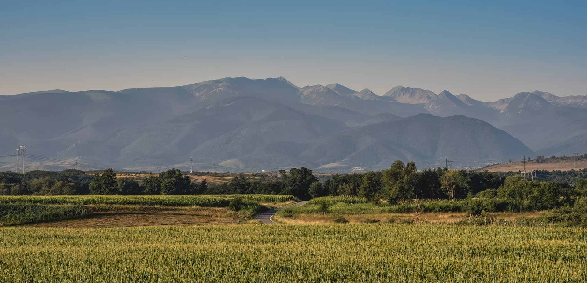 Mountain landscape with hazy peaks over green fields and a winding road in Retezat area.