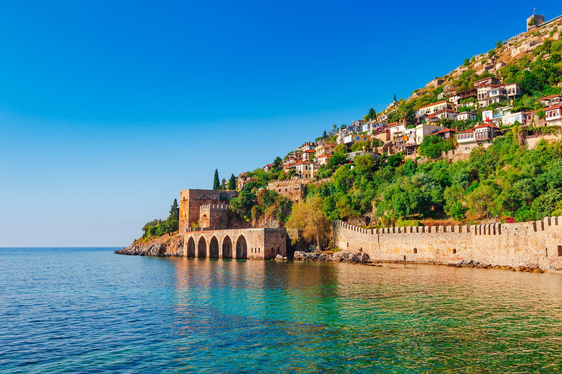 Historic stone fortress walls meeting turquoise water below houses climbing a lush green hill in Antalya.