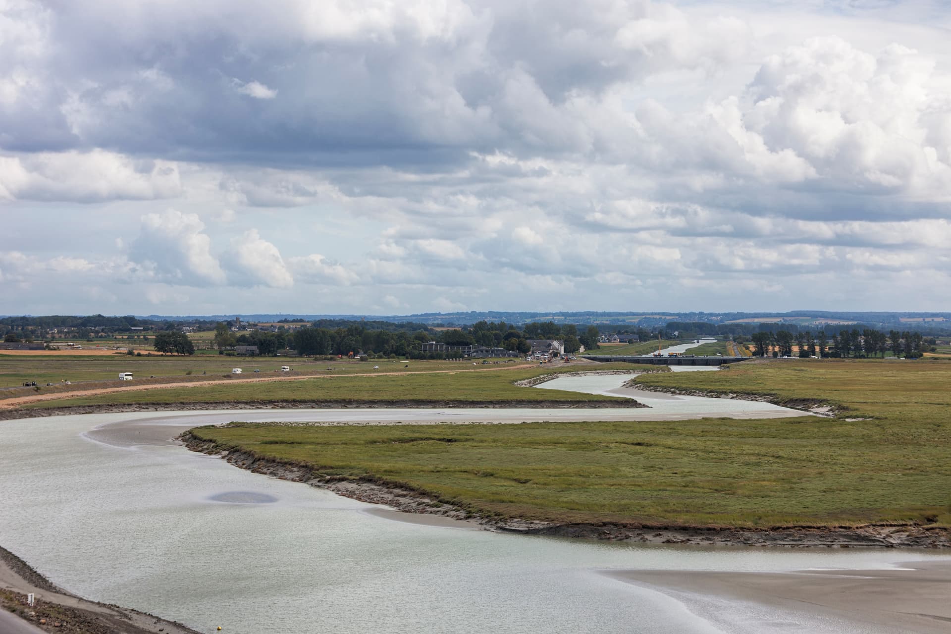 Winding river channel through marshland under cloudy sky near Mont Saint-Michel.