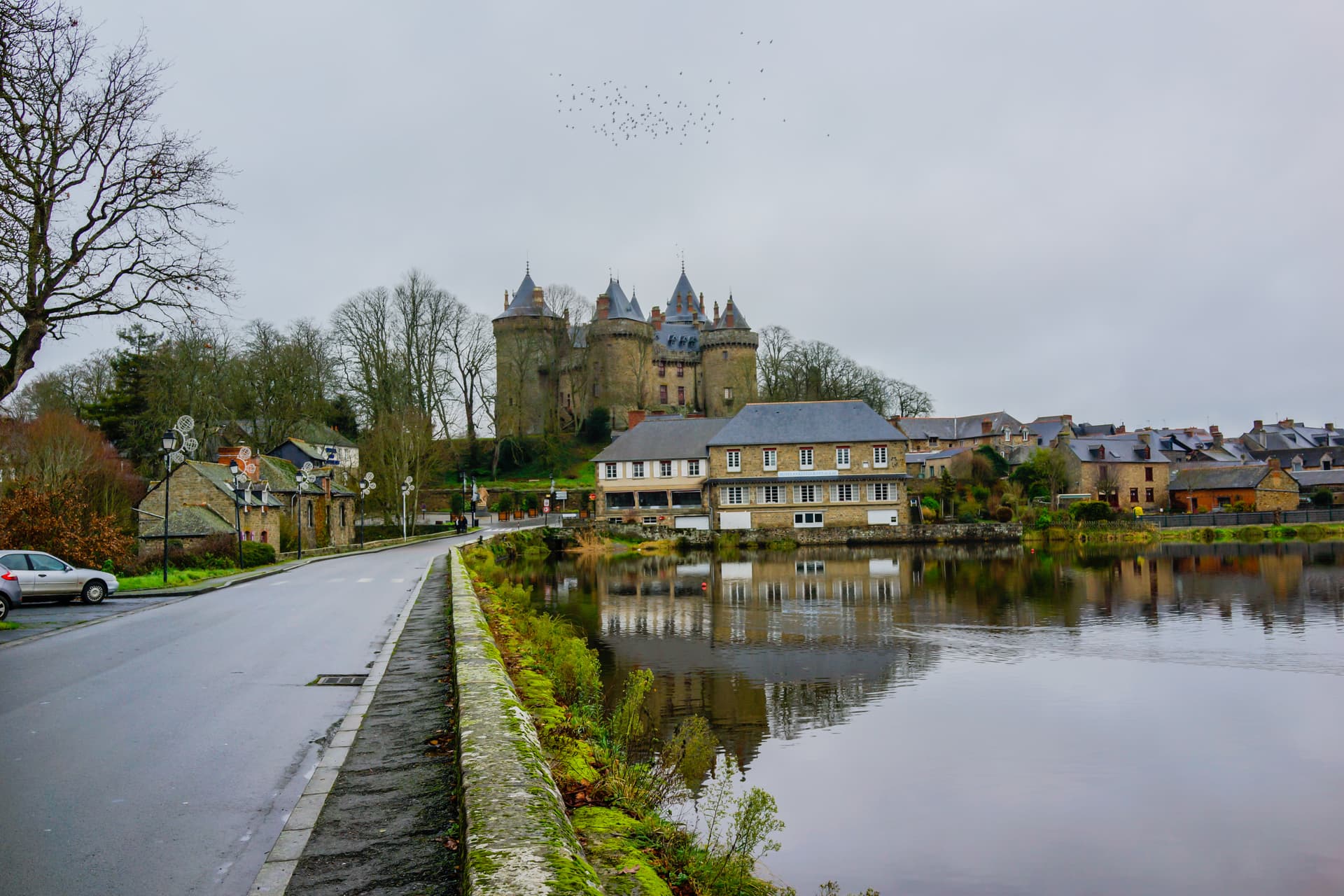 Château de Dol-de-Bretagne reflected in calm water beside a road with stone buildings.