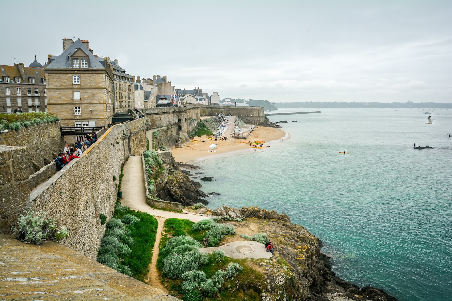 Coastal city Saint-Malo with stone ramparts, beach, and turquoise sea under an overcast sky.