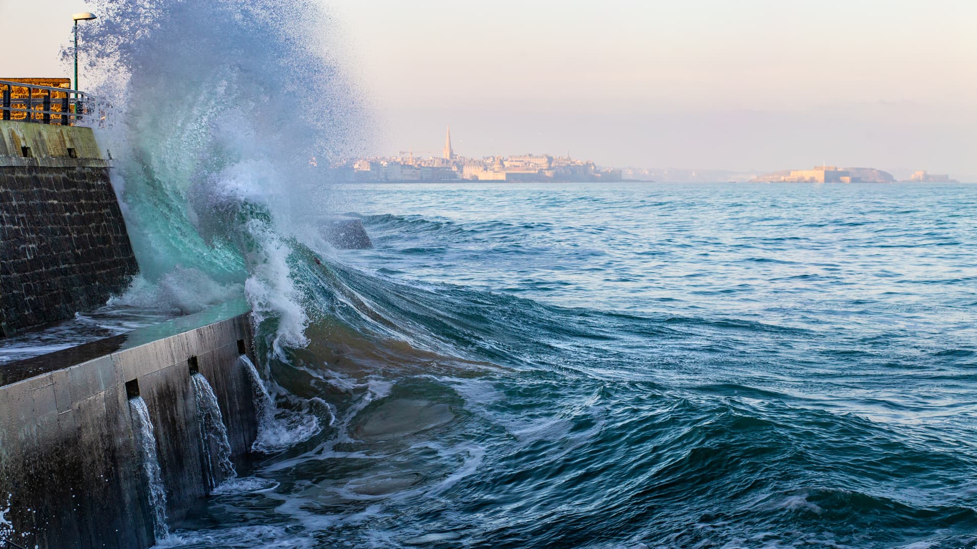 Big wave crashing against sea wall with Saint-Malo skyline in background.