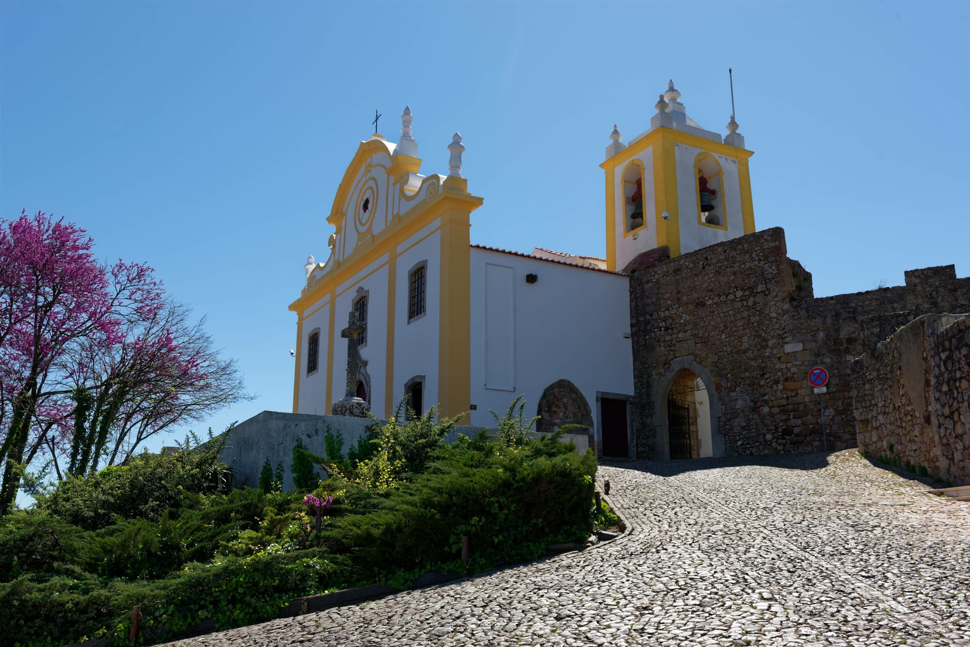 White and yellow church with bell tower next to stone walls on cobblestone street in Santiago do Cacém, Portugal.