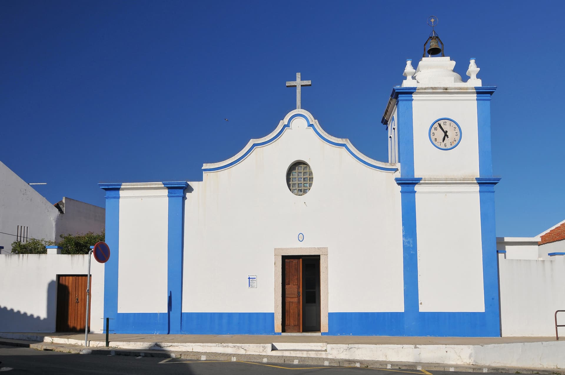 White and blue church facade with clock tower under clear blue sky in Vila Nova de Milfontes, Portugal.