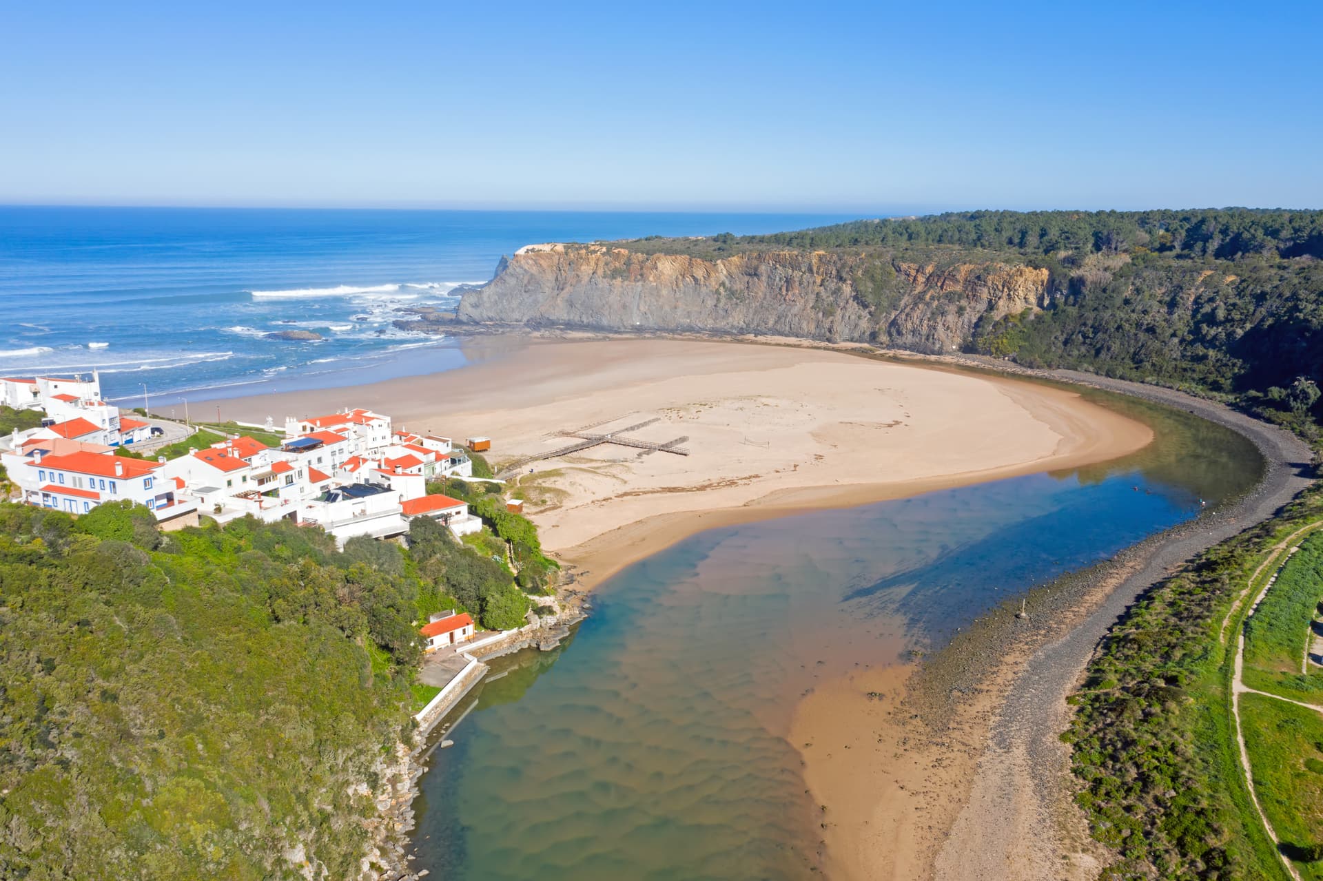 Aerial view of Praia de Odeceixe with white houses, river meeting the sea, and cliffs in Portugal.