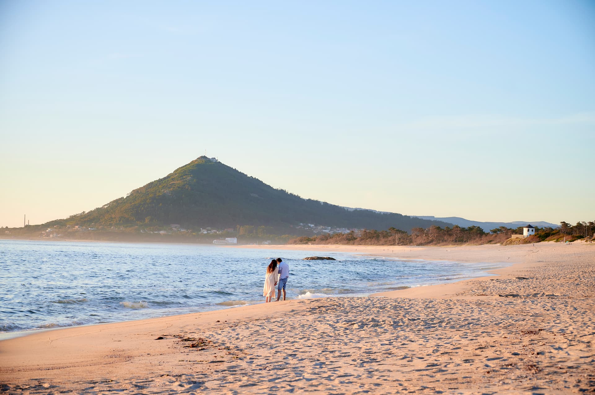 Woman in white dress and man on beach at Moledo, Portugal, with mountain background.