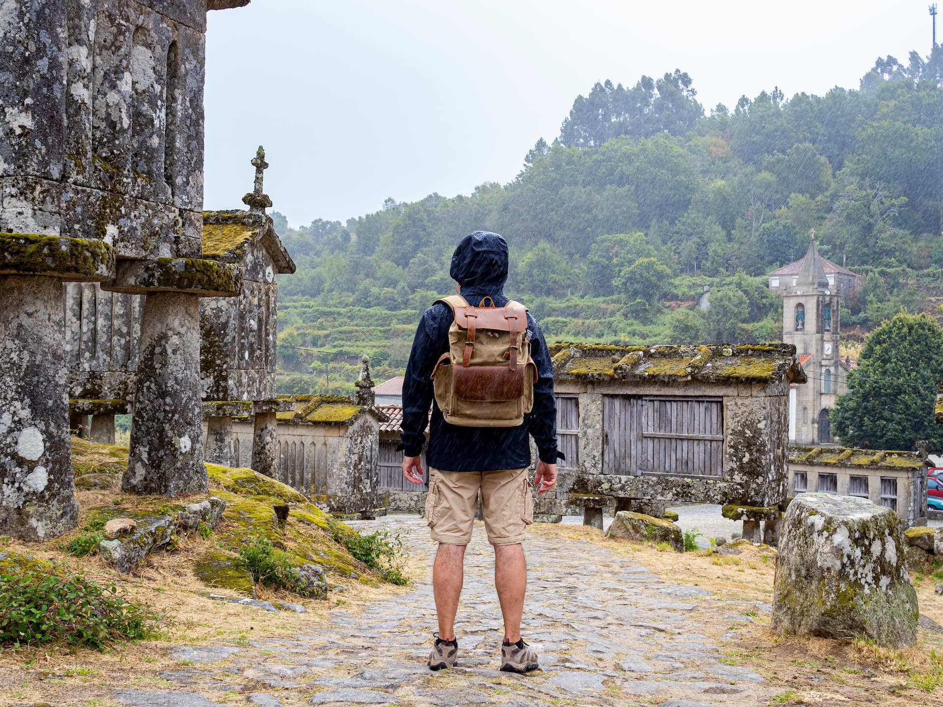 Hiker with backpack looking at historic stone granaries in Lindoso, Portugal as it rains.