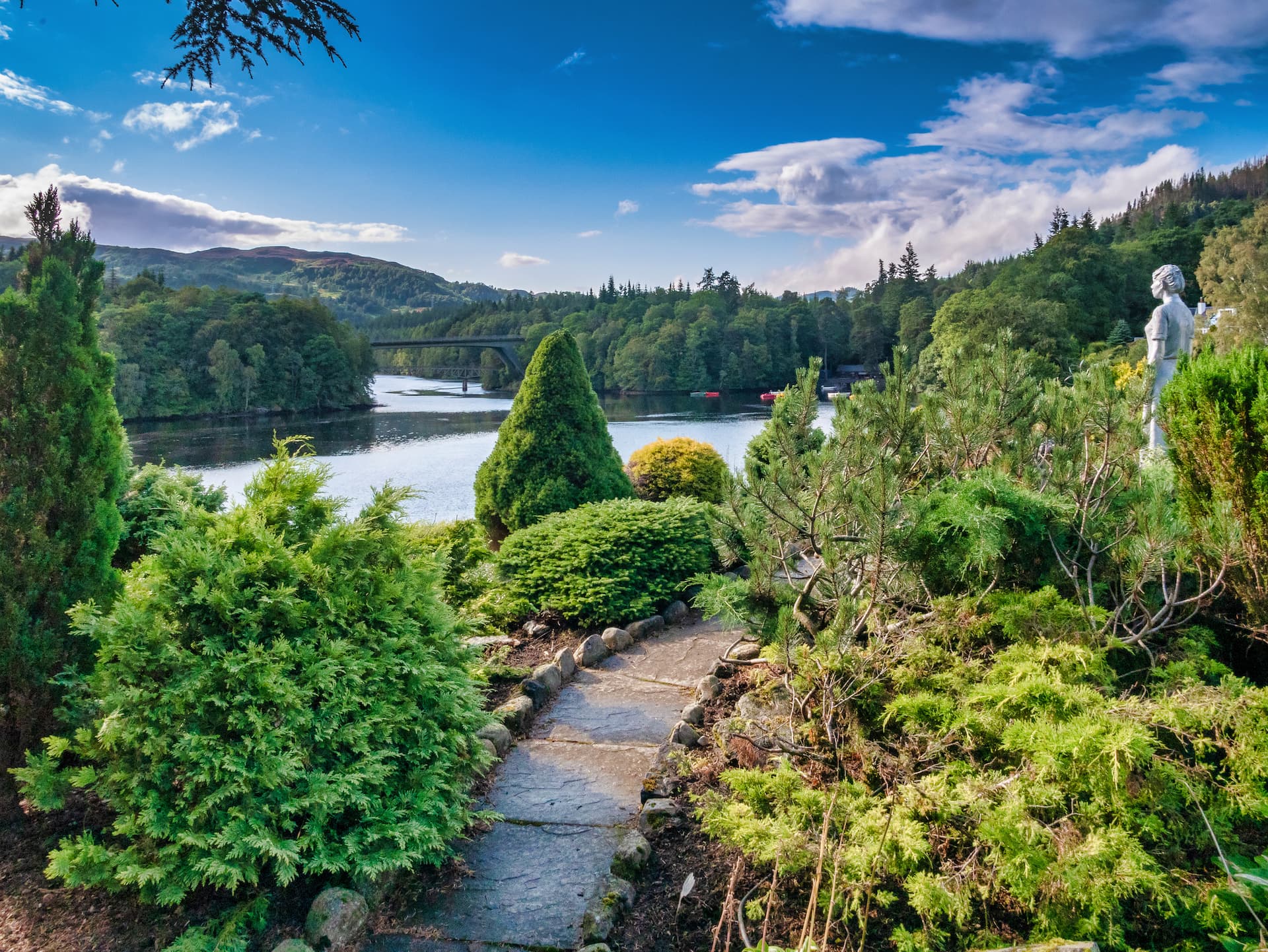 Stone path through lush garden overlooking river, bridge, and forested hills in Pitlochry.