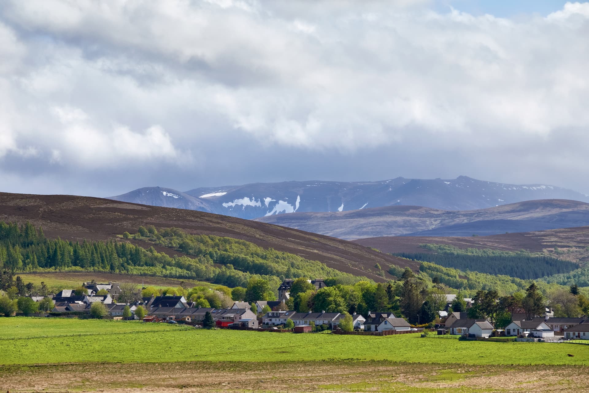 Village nestled in green valley with snow-capped mountains under cloudy sky, Tomintoul.