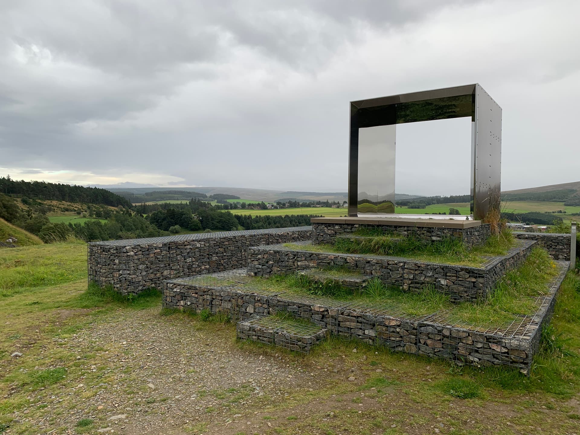 Modern art installation with gabion walls overlooking green rolling hills under a cloudy sky.