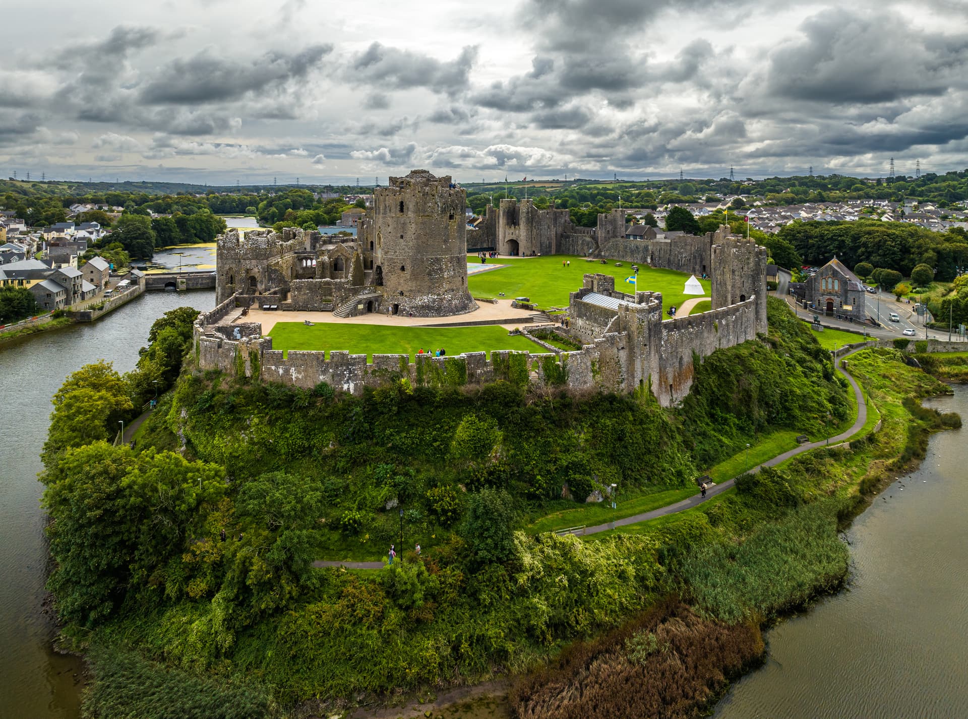 Stone castle ruins above a river with lush green banks under a cloudy sky, Pembroke.