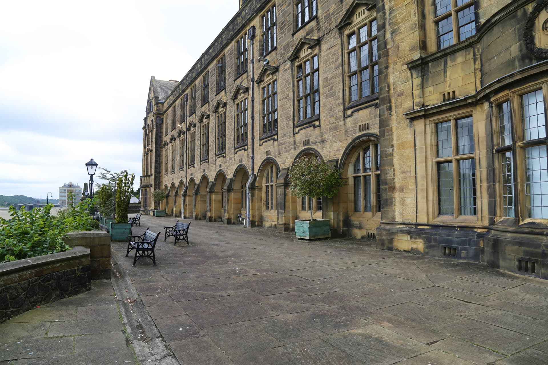 Stone building exterior with arched colonnade, benches, and paved terrace, Bangor University.