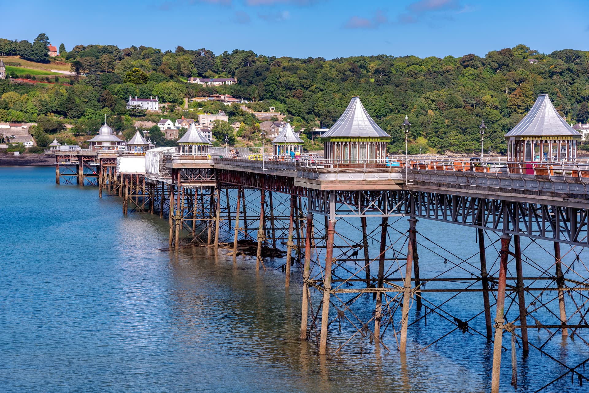 Bangor Pier structure over blue water with wooded hillside and houses in background.