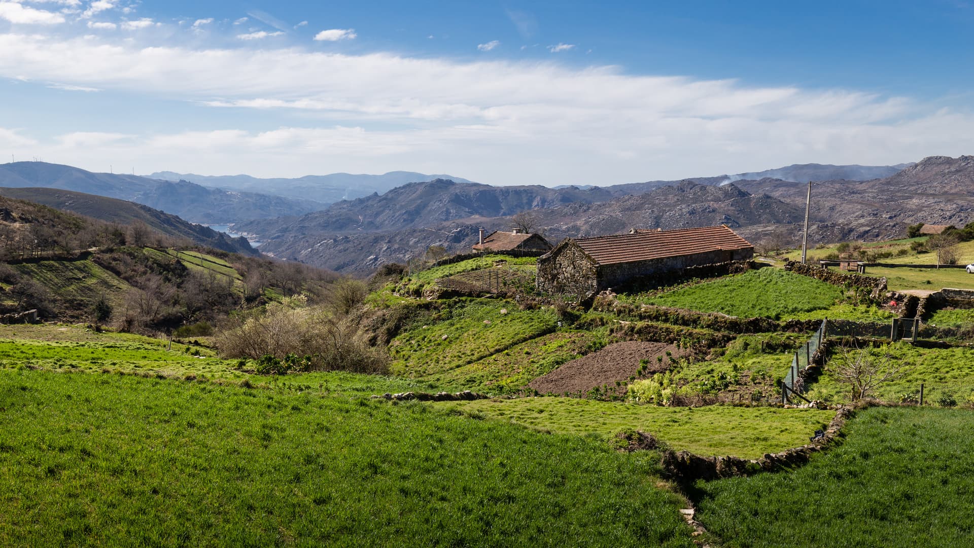 Rural landscape with stone houses, green terraces, and mountains under a blue sky at Pitões das Júnias, Portugal.