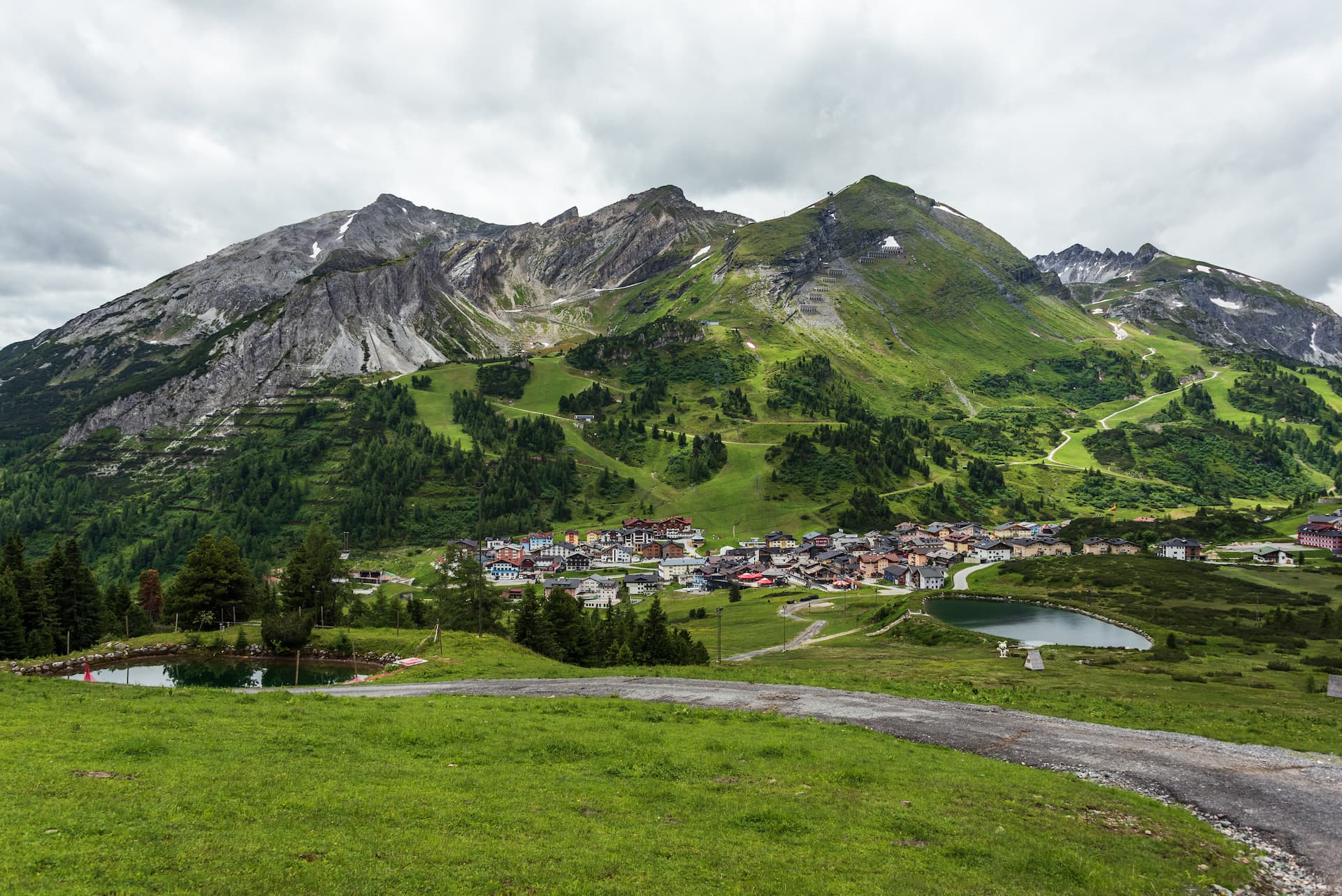 Alpine village nestled below green mountains with small ponds under cloudy sky in Obertauern.