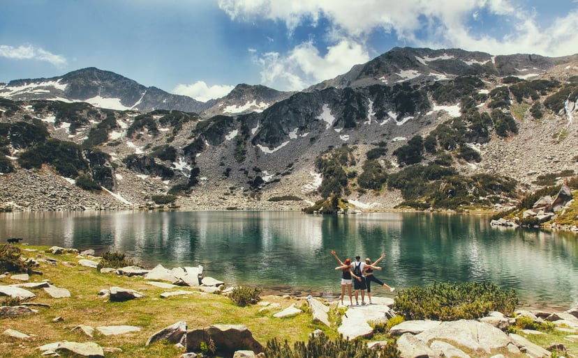 Hikers celebrating by clear alpine lake with rocky mountains and snow patches, Pirin Lakes