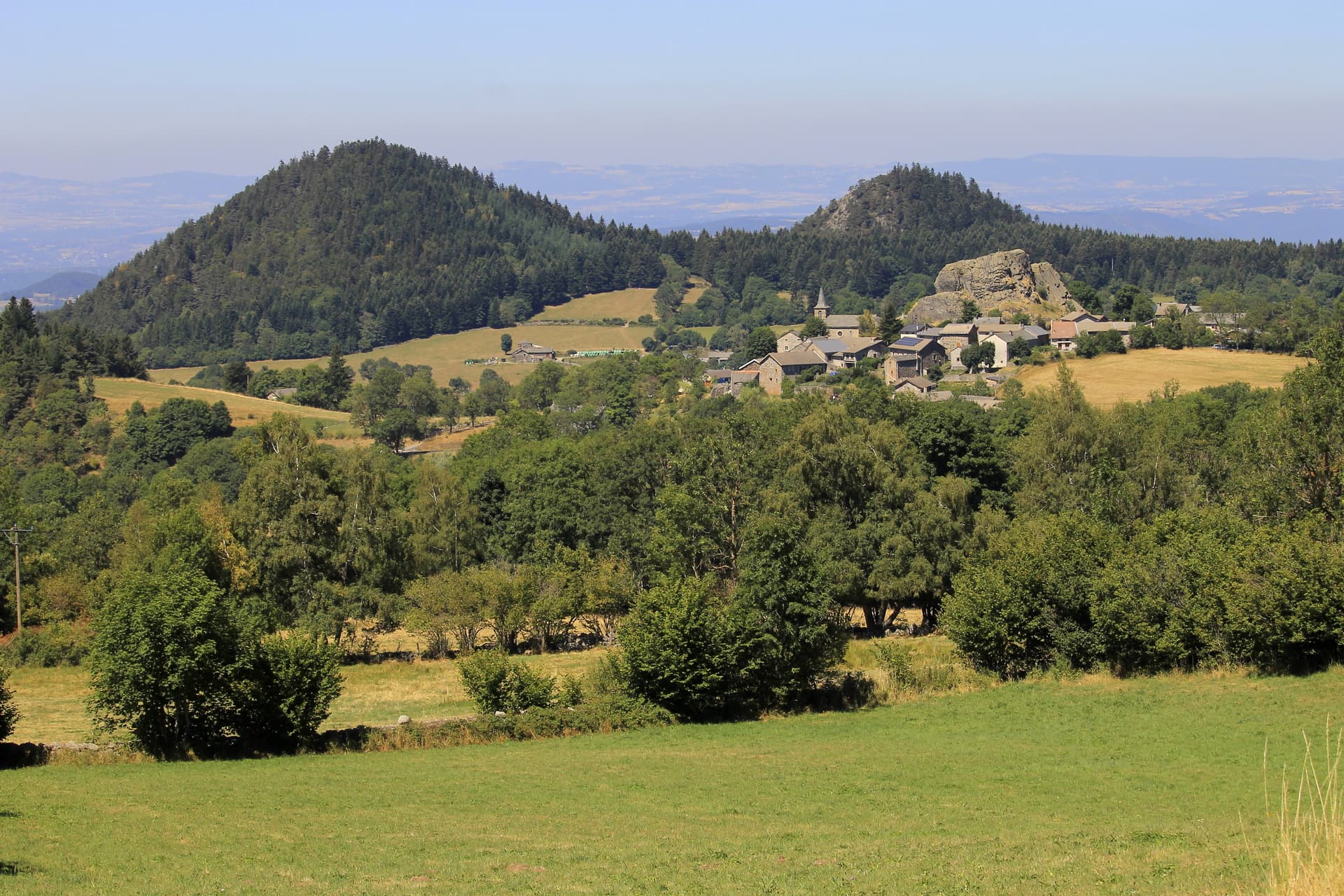 Village nestled in green hills with forested mountains in the background, Queyrières, France.