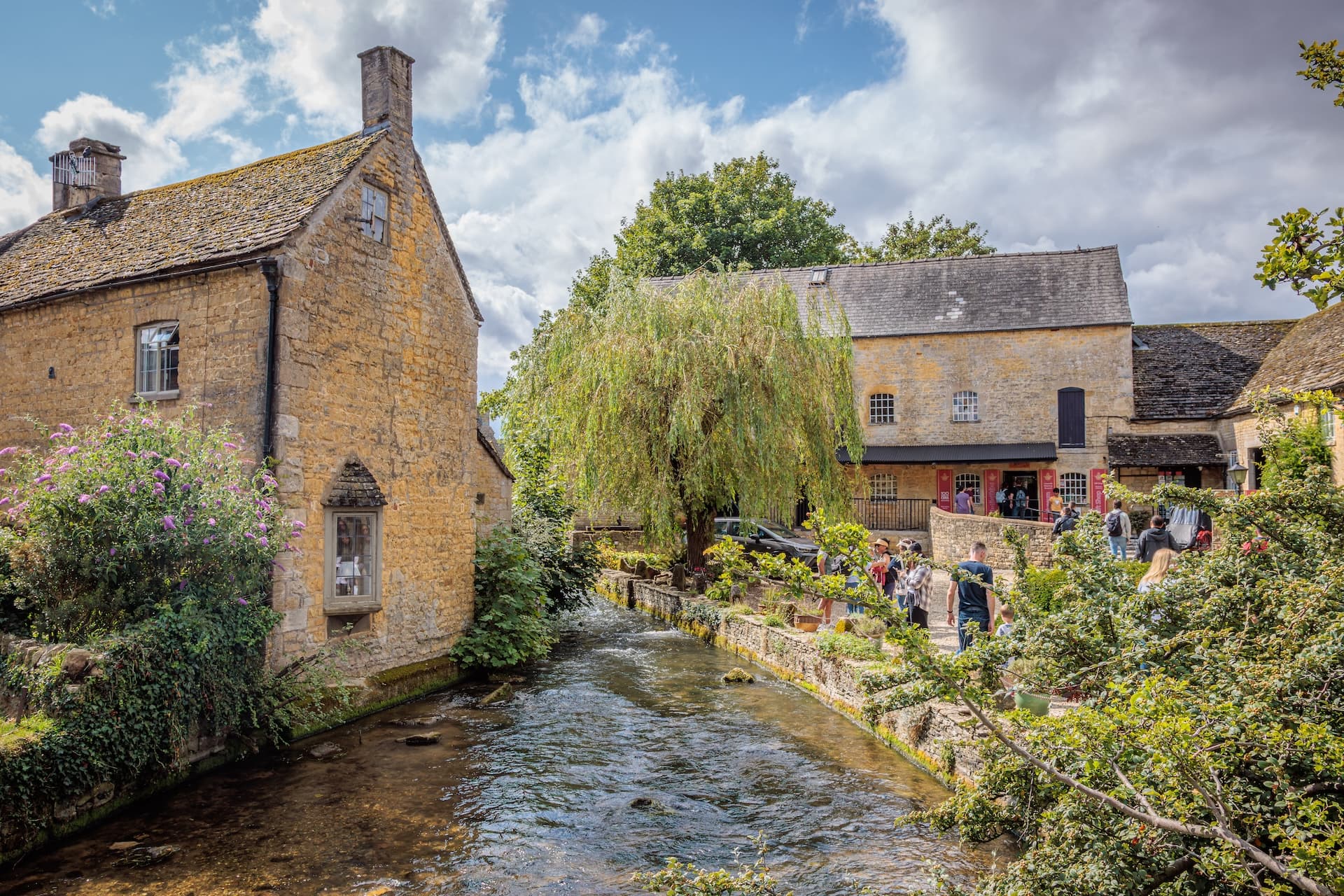 Stone buildings flank a stream with tourists walking near a weeping willow in Bourton-on-the-Water, England.