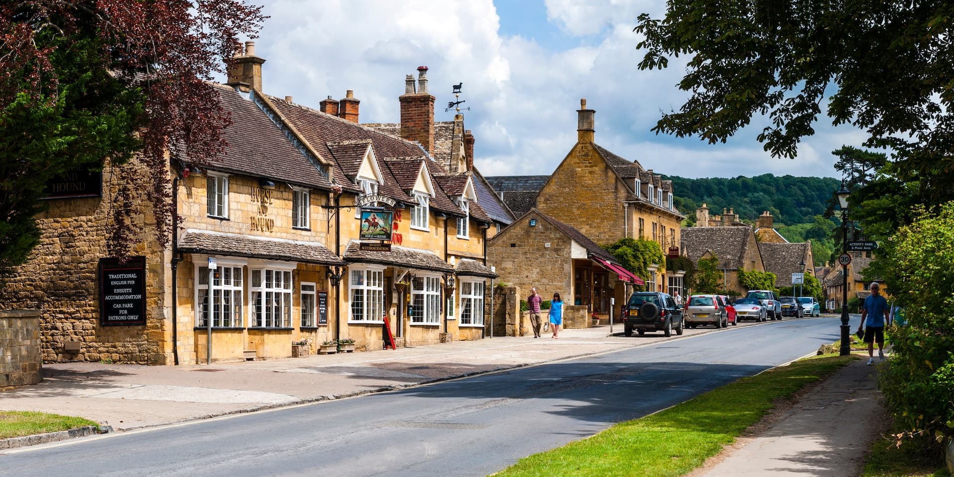 Cotswold village street with stone buildings, pub sign, and green hills in Gloucestershire.