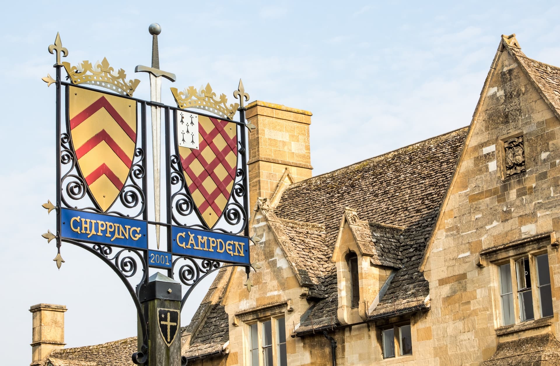 Chipping Campden sign with heraldic shields next to historic Cotswold stone building.