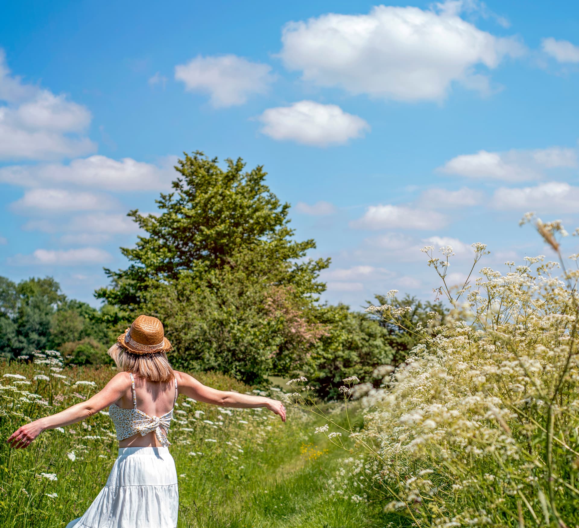 Woman in straw hat walking through tall grass and white wildflowers under blue sky.