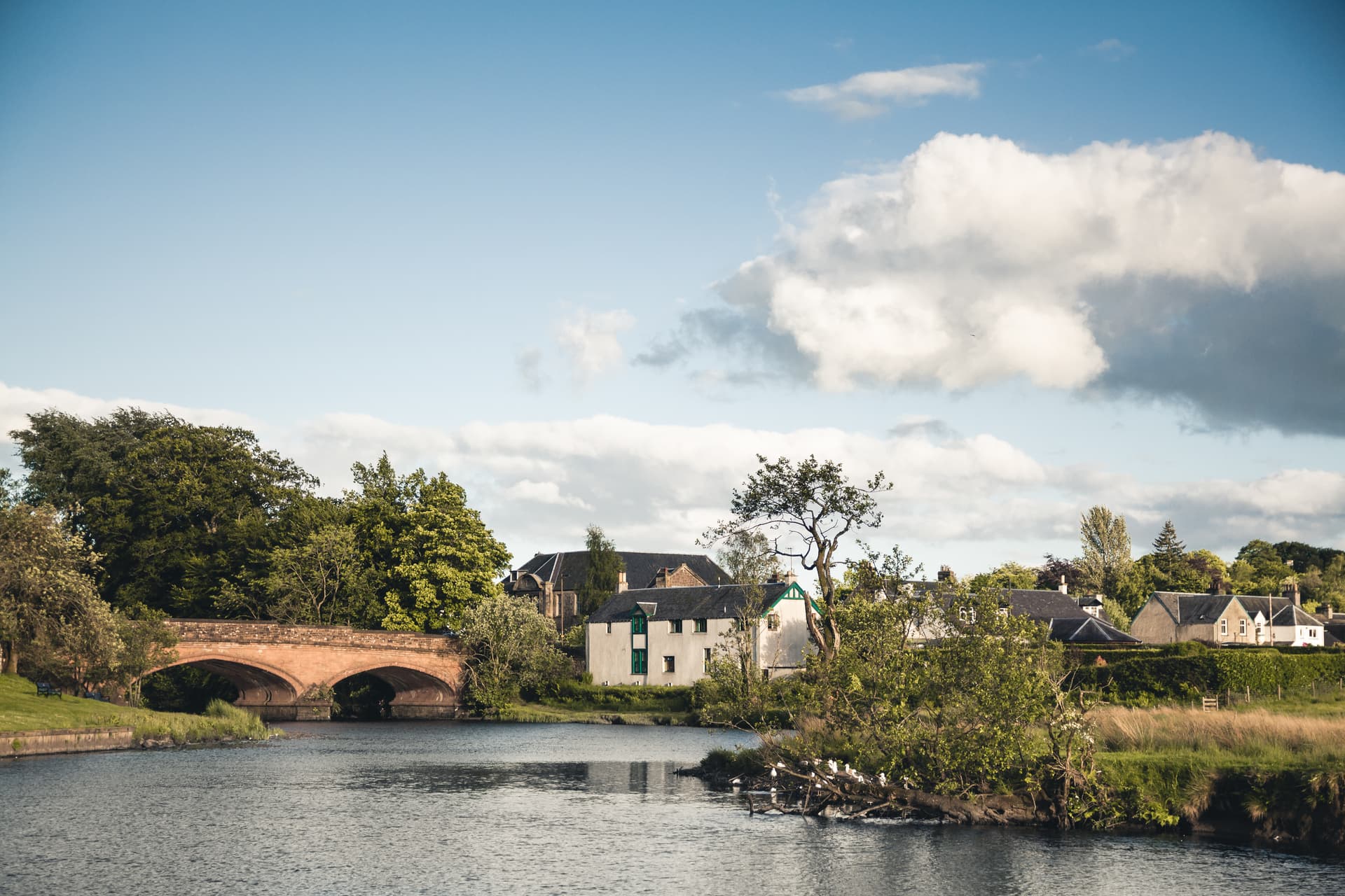 Stone arch bridge over river near white houses in Callander under a partly cloudy blue sky.