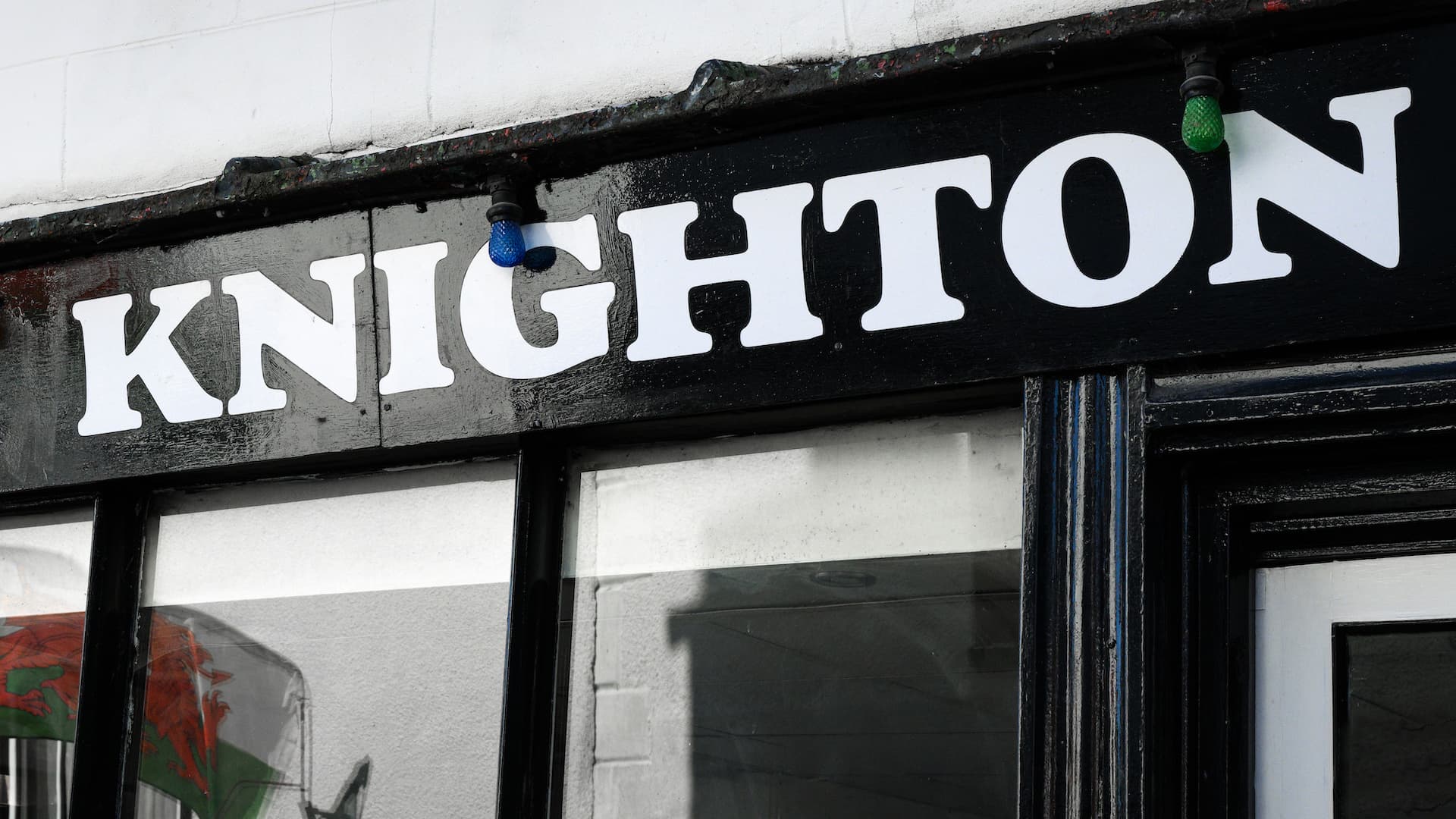 Black sign with white "KNIGHTON" lettering and colored light bulbs above shop windows.