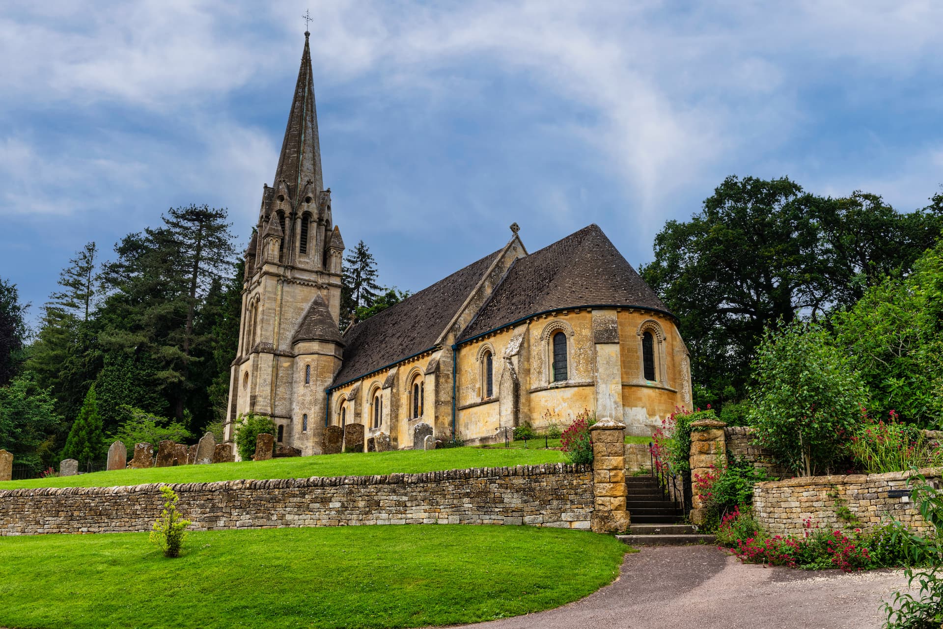 Stone church with tall spire, graveyard, and dry stone wall in Moreton-In-Marsh.