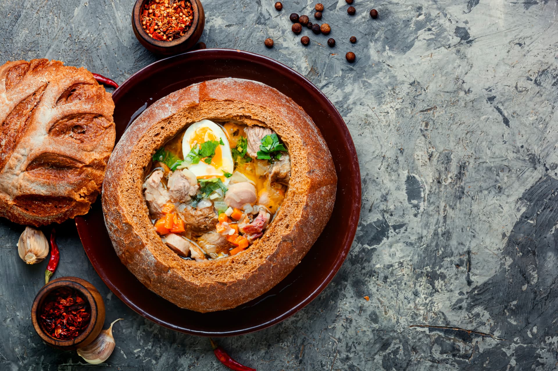 Stew served in a round bread bowl with hard-boiled egg, on a dark textured surface with spices.