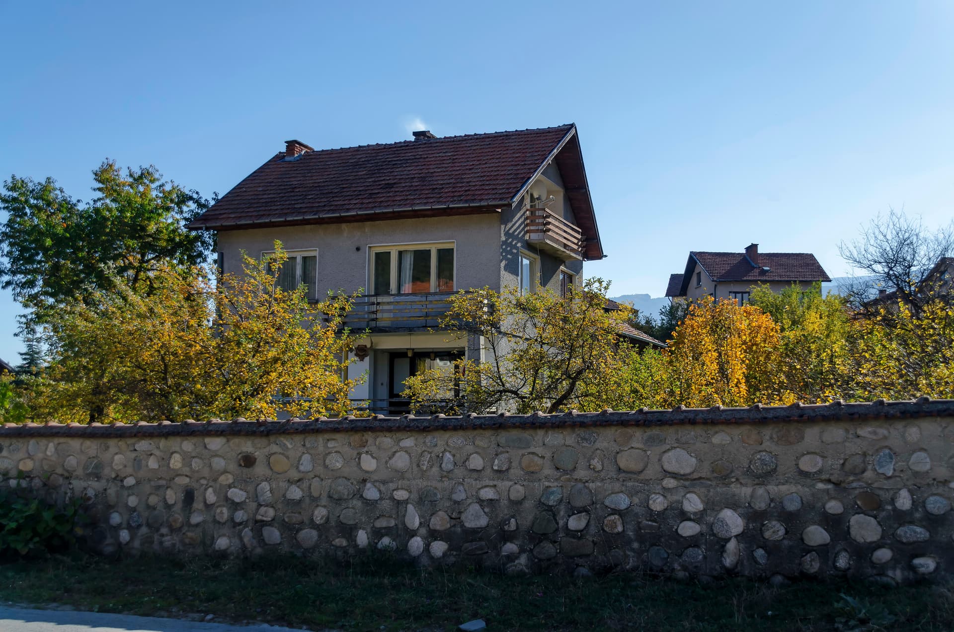 Two-story house with autumn trees behind a rounded stone wall in Govedartsi.