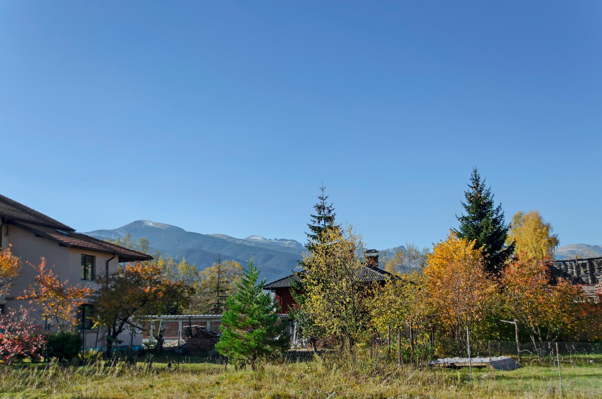 Houses in Govedartsi with autumn trees and snow-dusted mountains under a clear blue sky.
