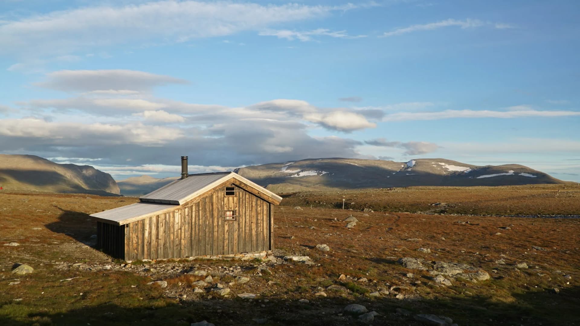 Wooden hiking hut near Gjendesheim, Norway, with mountains and snow patches under a cloudy sky.