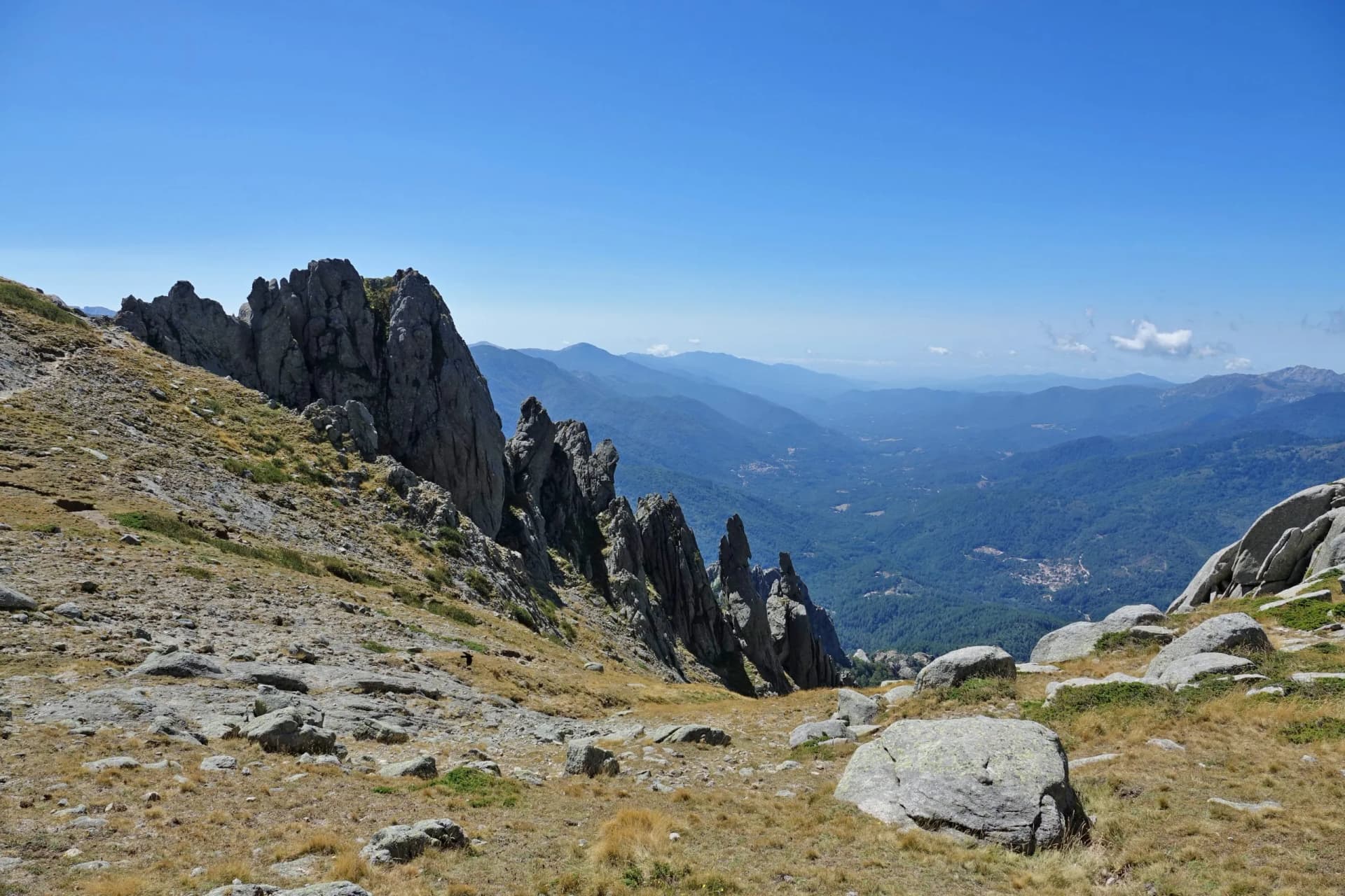 Hiking near jagged rock formations overlooking a vast, forested valley under a clear blue sky in Corsica.