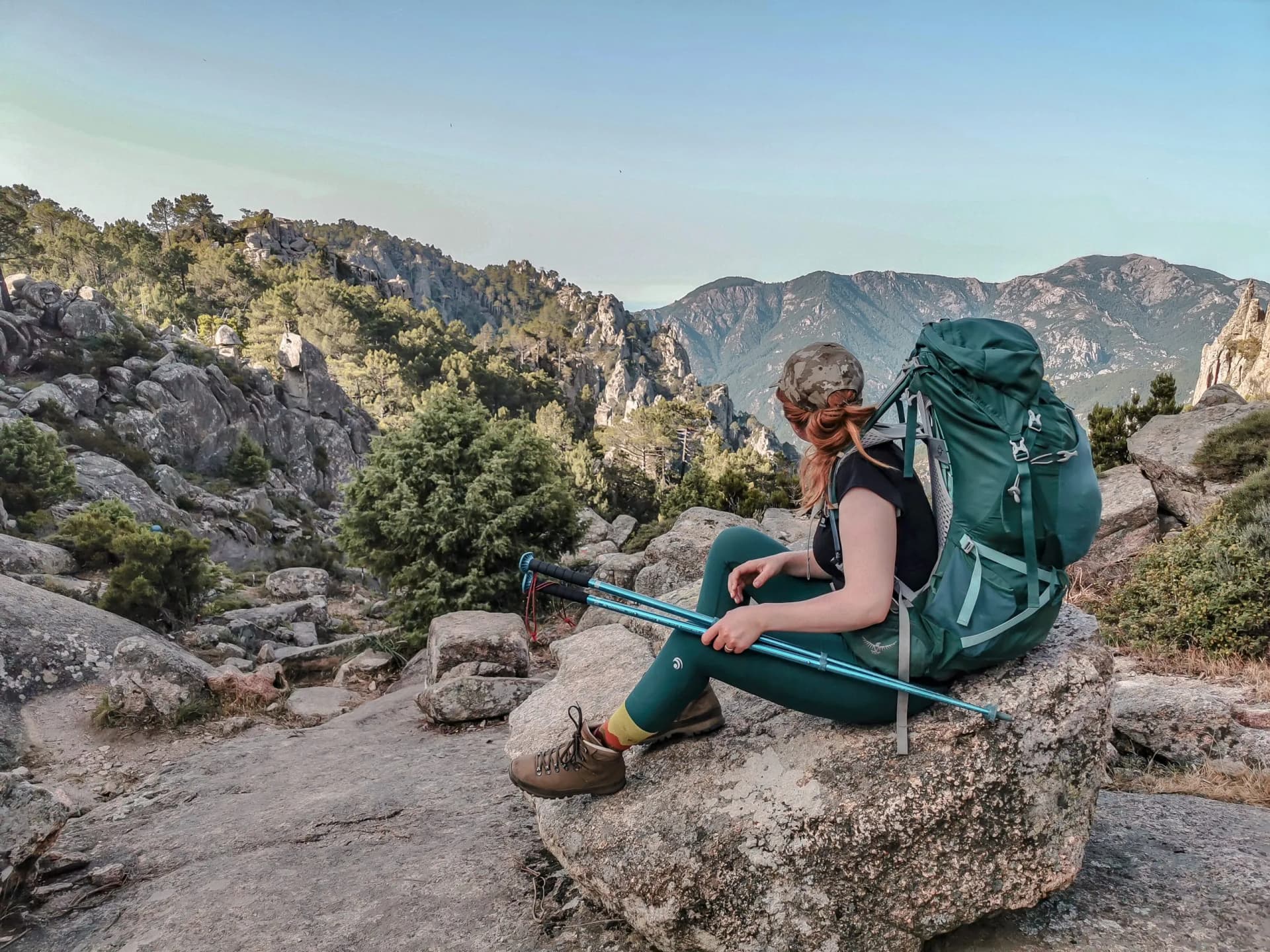 Hiker resting with heavy backpack and trekking poles overlooking mountains on the GR20 trail in Corsica.
