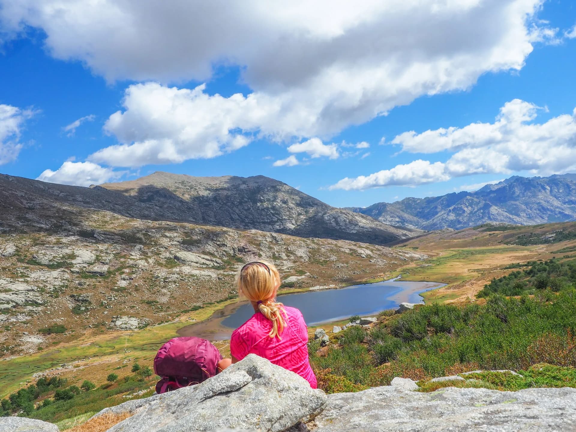 Hiker resting overlooking Lac de Nino, Corsica, with mountains and blue sky.