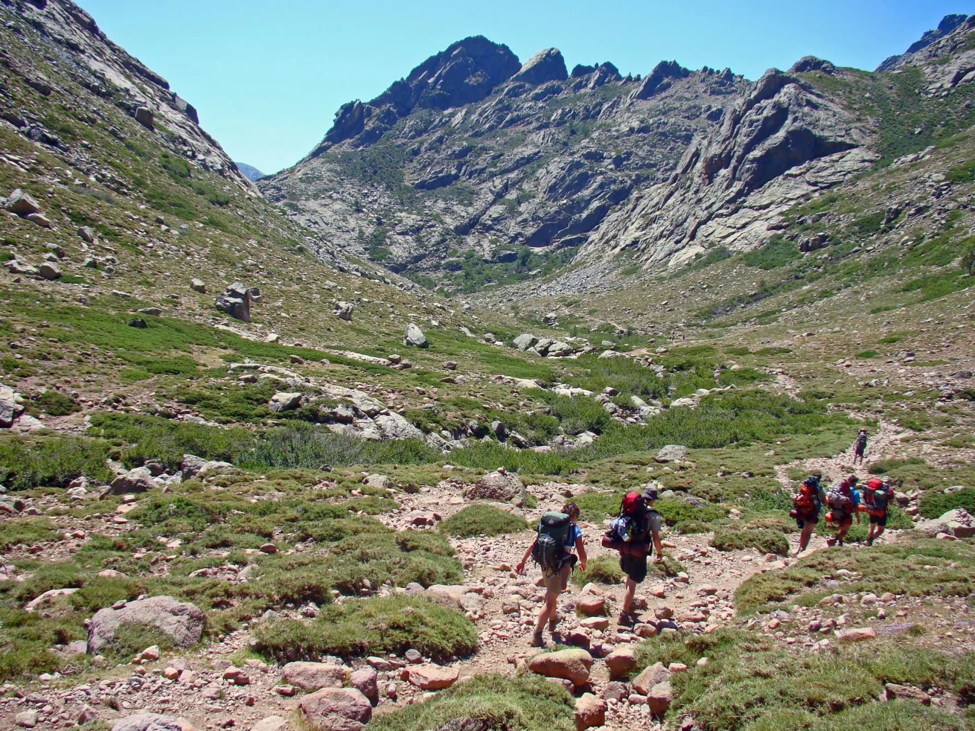 Hikers with backpacks trekking through a rocky, grassy valley toward steep mountains under a clear blue sky.