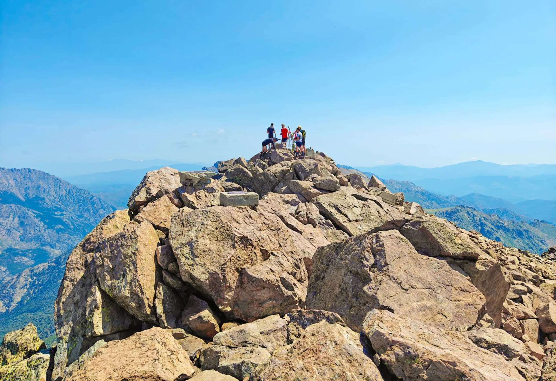 Hikers on Monte Cinto peak, Corsica, with views of blue mountain ranges under a clear sky.