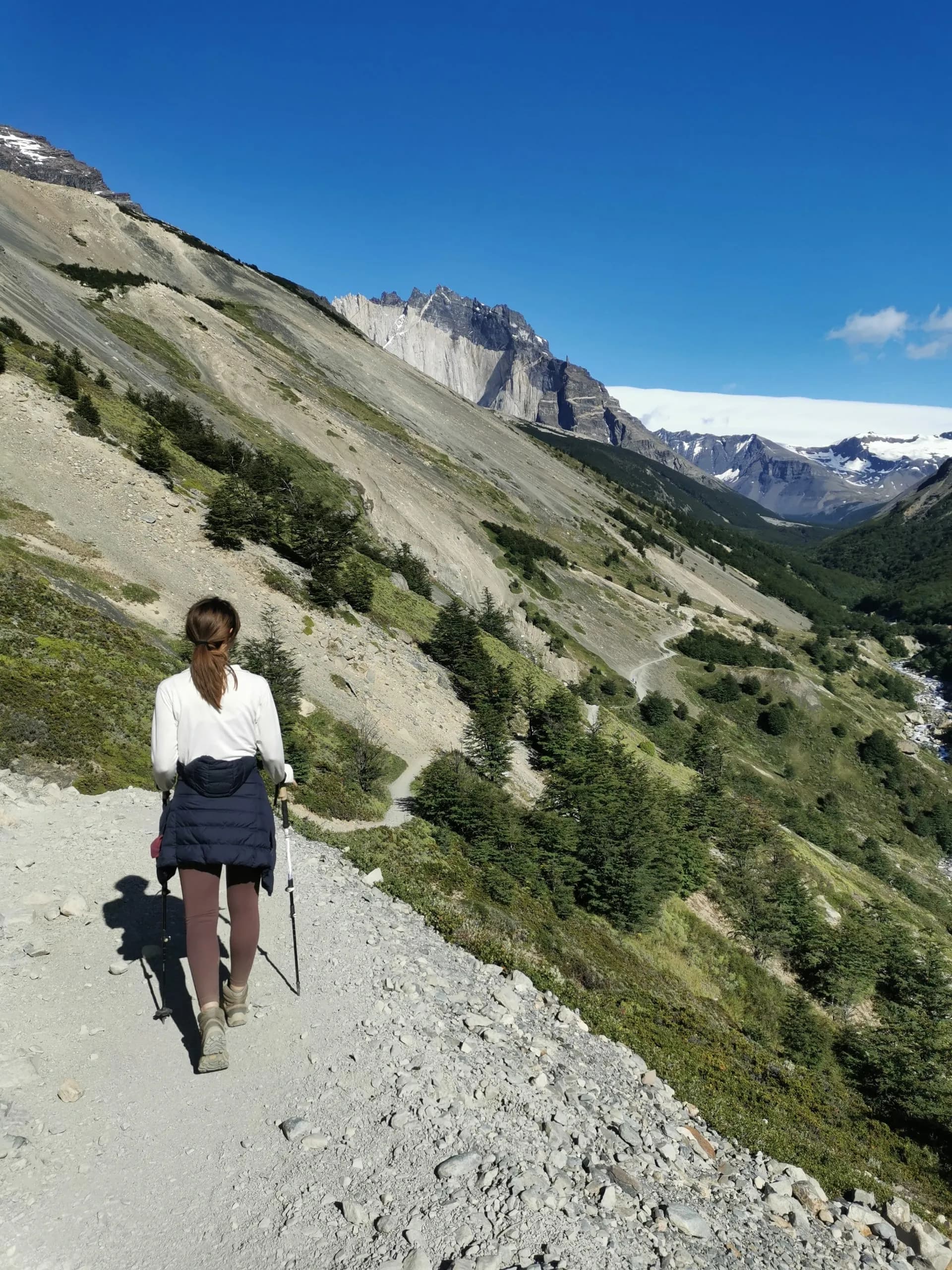 Hiker with trekking poles walking on rocky mountain trail toward jagged, snow-capped peaks under blue sky.